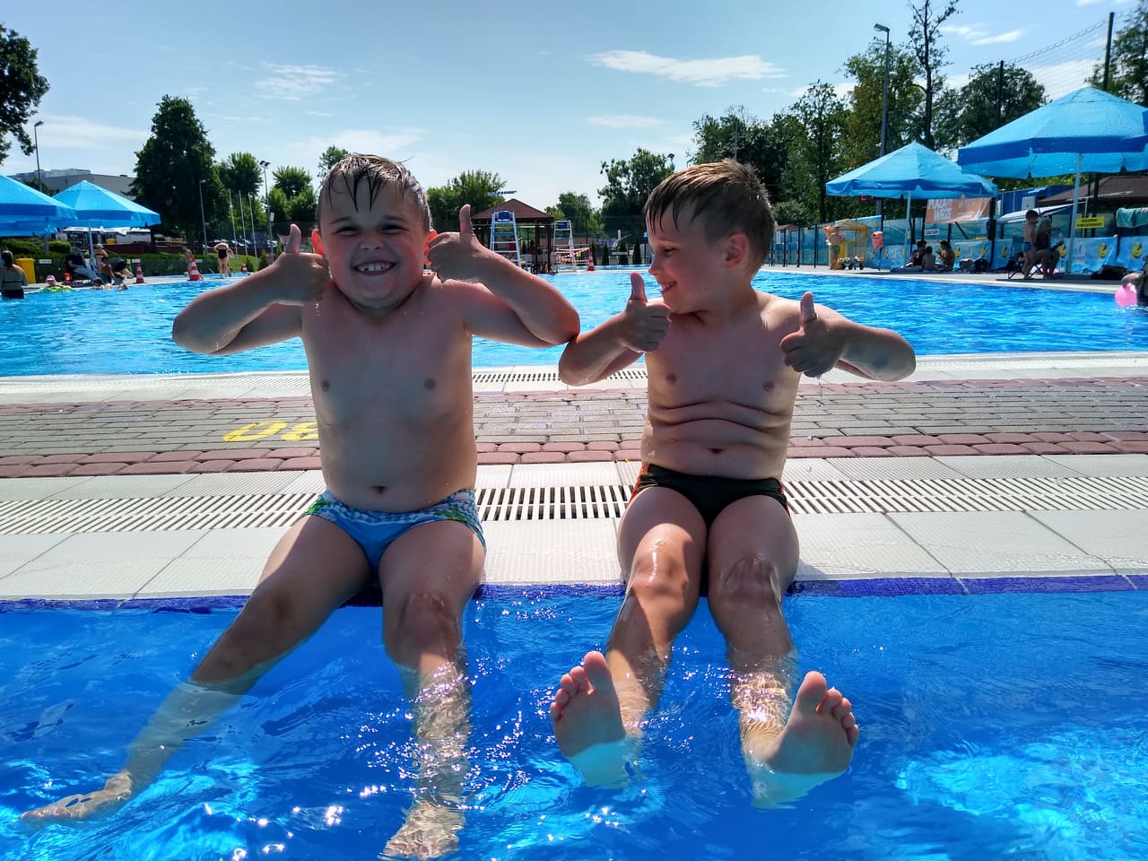 chłopcy siedzą na brzegu basenu, uśmiechają się i pokazują kciuki w górę